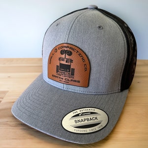 May include: Gray and black baseball cap with a brown leather patch featuring a robot and the text "WALL-E Compacting Co. Waste Allocation Load Lifter Earth Class est. 2028". The cap also has a gold label with the text "The Authentic Yupoong Snapback Yupoong Since 1974".