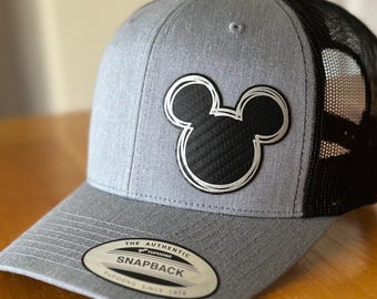 May include: A gray and black baseball cap with a black Mickey Mouse head logo. The cap has a mesh back and a snap closure. The label on the front reads "The Authentic of Trucker Snapback Trucker Since 1974."