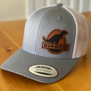 May include: Gray and white baseball cap with a brown leather patch featuring a black dinosaur silhouette and the text "Dinoco". The patch is on the front of the cap. The cap has a white mesh back and a black "The Authentic Yupoong Snapback Yupoong Since 1974" label on the underside of the brim.