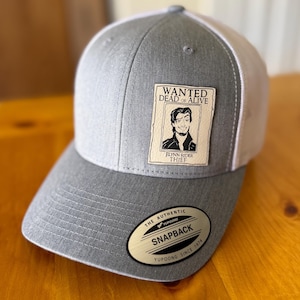 May include: Gray and white Snapback hat with a curved brim. The hat features a patch with a "Wanted Dead or Alive" poster of Flynn Rider, and the text "Flynn Rider Thief". The hat has a Yupoong Snapback label.