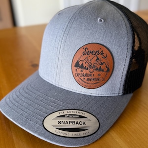 May include: Gray and black trucker hat with a curved brim. The hat features a circular leather patch with the text "Sven's Exploration Adventure Co." and a mountain and deer graphic. The hat also has a "Snapback" label.