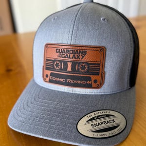 May include: Gray and black snapback hat featuring a brown leather patch with the text "GUARDIANS OF THE GALAXY" and "Cosmic Rewind" along with a cassette tape design. The hat has a curved brim and a "Yupoong" tag.
