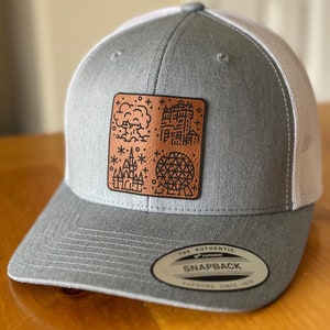 May include: A gray and white trucker hat with a brown leather patch featuring a stylized illustration of a castle, a sphere, and a cloud with the text "The Most Magical Place on Earth". The hat has a snap closure and the text "The Authentic Snapback Yupoong Since 1974" on the back.