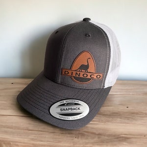 May include: A gray and white trucker hat with a brown leather patch featuring a dinosaur and the text "DINO CO". The hat has a black adjustable snap closure with the text "THE AUTHENTIC YUPOONG SNAPBACK YUPOONG SINCE 1974".