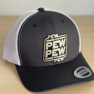 May include: A black and white trucker hat with a gold patch that says "PEW PEW PEW PEW" in a retro video game style font. The hat has a mesh back and a snap closure.