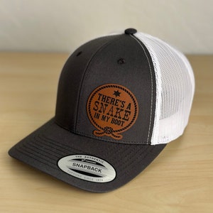 May include: A gray and white trucker hat with a brown leather patch that says "There's a Snake in My Boot". The hat has a mesh back and a snap closure.