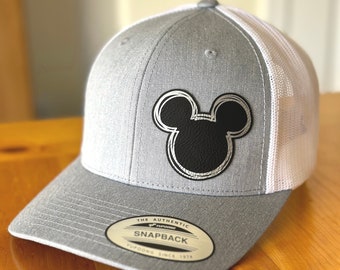 May include: A gray and white baseball cap with a black Mickey Mouse silhouette on the front. The cap has a mesh back and a snap closure. The label on the cap reads "The Authentic Yupoong Snapback Since 1974."