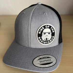 May include: A gray and black trucker hat with a black and white patch that says "Rise of the Resistance" with a Stormtrooper helmet graphic. The hat has a black mesh back and a gray front. The hat has a label that says "The Authentic Yupoong Snapback Yupoong Since 1974."