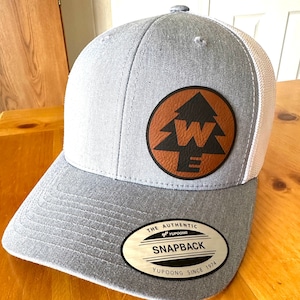 May include: Gray and white mesh trucker hat with a brown leather patch featuring a black silhouette of a pine tree with a white letter "W" inside. The patch has the text "THE AUTHENTIC SNAPBACK YUPOONG SINCE 1974" on it.