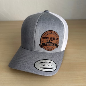 May include: A gray and white baseball cap with a brown leather patch that says "Mos Eisley Cantina" with the text "Live Music Hot Food No Droids". The patch has a small image of a sombrero. The cap has a white mesh back and a black "Yupoong Snapback" label.