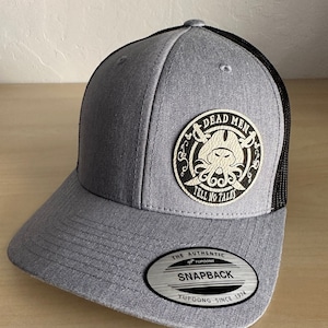 May include: A gray and black trucker hat with a black mesh back. The front of the hat features a white leather patch with a skull and crossbones design and the text "Dead Men Tell No Tales". The hat also has a silver label that says "The Authentic Yupoong Snapback Yupoong Since 1974".