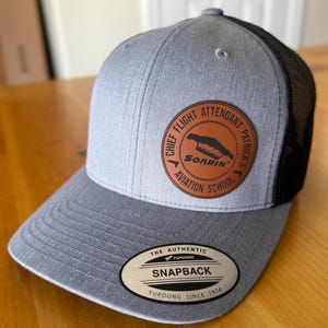 May include: Gray Snapback hat with a black mesh back. The hat features a circular leather patch with the text "CHIEF FLIGHT ATTENDANT PATRICK'S SOARIN' AVIATION SCHOOL". The brim has a "SNAPBACK" label.