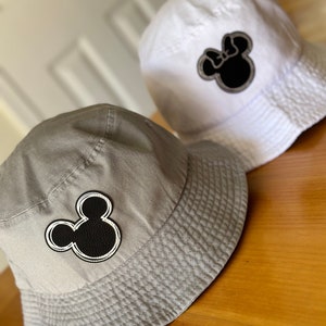 May include: Two bucket hats with black and white Mickey Mouse ears embroidered on them. One hat is gray and the other is white.