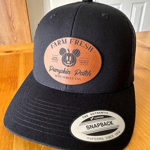 May include: Black and brown baseball cap with a leather patch that says "Farm Fresh Pumpkin Patch Main Street USA" and a Mickey Mouse head with a pumpkin face. The patch has a brown leather background and white stitching. The cap has a black mesh back and a black snap closure with a silver label that says "The Authentic YUPOONG Snapback YUPOONG Since 1974".