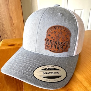 May include: A gray and white mesh baseball cap with a brown leather patch that says "Sven & Olaf's Reindeer Farm Est. 2013". The cap also has a silver oval patch that says "The Authentic YUPOONG Snapback YUPOONG Since 1974".