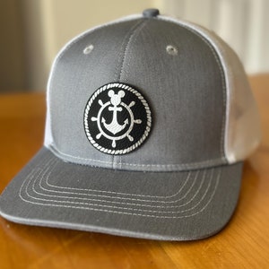 May include: A gray and white baseball cap with a black leather patch featuring a white anchor and Mickey Mouse ears. The patch is surrounded by a white rope design.