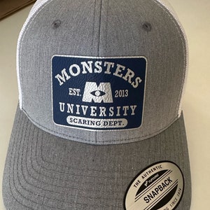 May include: Gray and white trucker hat with a blue patch that says "Monsters University Scaring Dept." and a white logo of the Monsters Inc. eye with the text "Est. 2013". The hat has a black and silver sticker that says "The Authentic YUPOONG Snapback YUPOONG Since 1974".