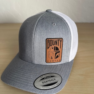 May include: Gray and white trucker hat with a brown leather patch that says "The Bounty Hunter" with a Mandalorian helmet graphic. The hat has a YUPOONG SNAPBACK label on the underside of the brim.