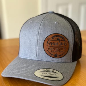 May include: A gray and black trucker hat with a brown leather patch that says "Captain Jack's Tortugan Spiced Rum" with a skull and crossbones logo. The hat has a black mesh back and a gray front with a white "The Authentic Snapback" label.