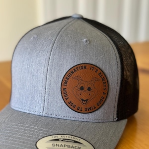 May include: A gray and black mesh trucker hat with a brown leather patch featuring a cartoon dragon and the text "It's always a good time to use your imagination."