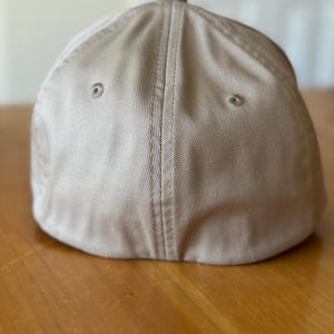K&ouml;nnte beinhalten: Eine beige Baseballkappe mit flachem Schirm und einer Naht, die die Mitte der Krone entlangl&auml;uft.
