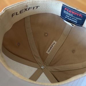 K&ouml;nnte beinhalten: Eine beige Baseballkappe mit einem wei&szlig;en Flexfit-Logo auf der Innenseite der Kappe. Die Kappe hat ein schwarzes Etikett mit dem Flexfit-Logo und dem Text "95% Polyester 5% Spandex Hergestellt in Vietnam YUPOONG" auf der R&uuml;ckseite.