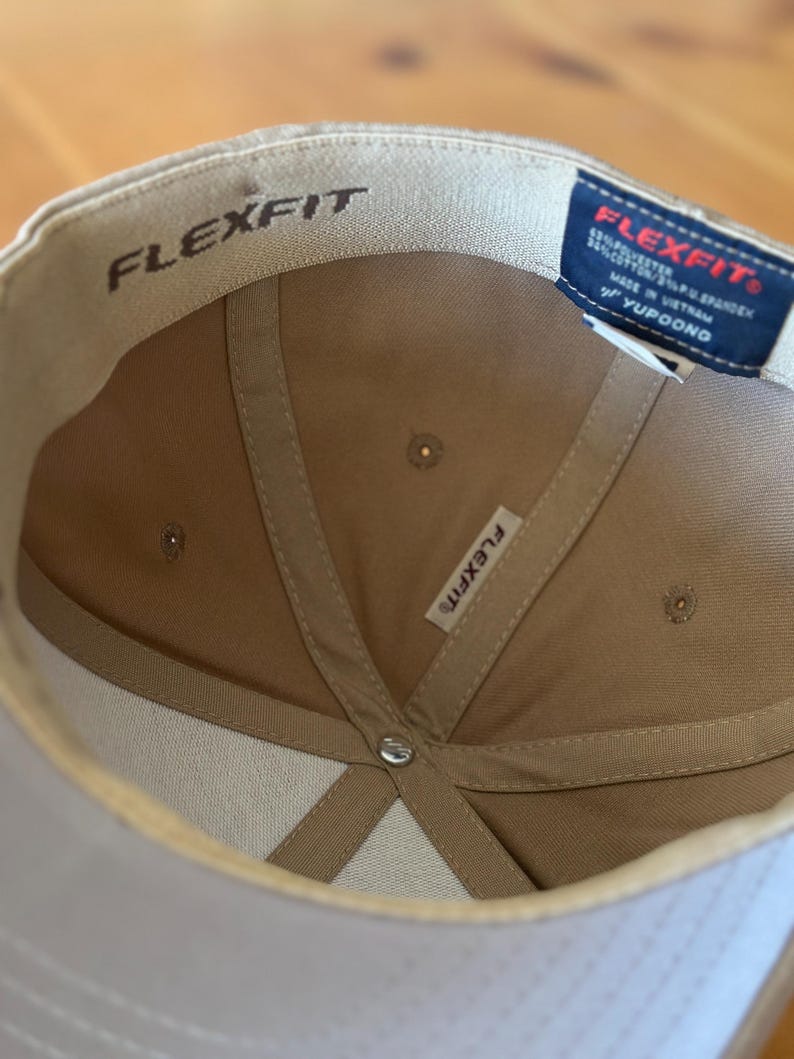 K&ouml;nnte beinhalten: Eine beige Baseballkappe mit einem wei&szlig;en Flexfit-Logo auf der Innenseite der Kappe. Die Kappe hat ein schwarzes Etikett mit dem Flexfit-Logo und dem Text "95% Polyester 5% Spandex Hergestellt in Vietnam YUPOONG" auf der R&uuml;ckseite.