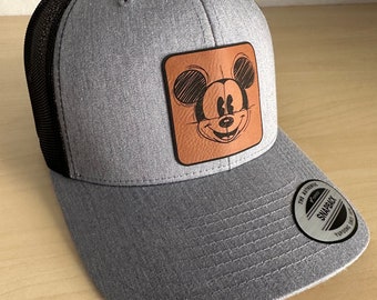 May include: A gray and black trucker hat with a brown leather patch featuring a cartoon image of Mickey Mouse. The hat has a mesh back and a snap closure.