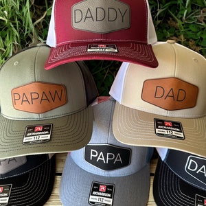 May include: Five different baseball caps with a leather patch on the front. The patches have the words "Daddy", "Papa", "Paw", "Dad", and "Papa" on them. The caps are all different colors, including red, green, beige, gray, and black.