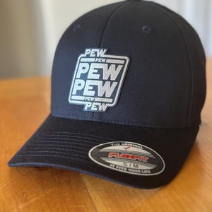 K&ouml;nnte beinhalten: Eine schwarze, geformte Baseballkappe mit einem silbernen gestickten Aufn&auml;her, auf dem in einem Quadrat "PEW PEW PEW" steht. Die Kappe hat ein Flexfit-Etikett auf der R&uuml;ckseite, auf dem "The Original Flexfit S/M It Fits Your Life." steht.