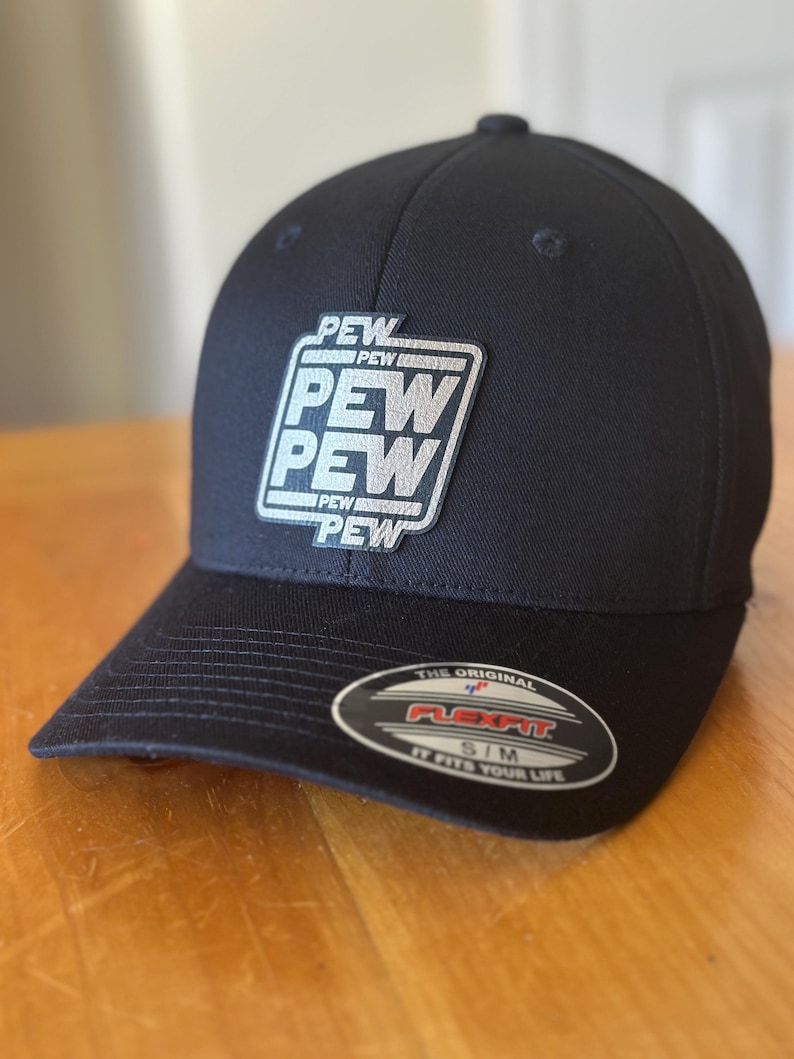 K&ouml;nnte beinhalten: Eine schwarze, geformte Baseballkappe mit einem silbernen gestickten Aufn&auml;her, auf dem in einem Quadrat "PEW PEW PEW" steht. Die Kappe hat ein Flexfit-Etikett auf der R&uuml;ckseite, auf dem "The Original Flexfit S/M It Fits Your Life." steht.