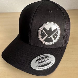 May include: Black and silver baseball cap with a logo that says "Strategic Homeland Intervention Enforcement Logistics Division" and a silver eagle. The cap has a black mesh back and a silver label that says "The Authentic Yupoong Snapback Yupoong Since 1974".