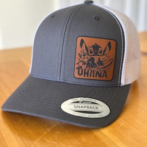 May include: A gray and white trucker hat with a brown leather patch featuring a cartoon Stitch character from Lilo & Stitch. The patch has the word "Ohana" written on it.