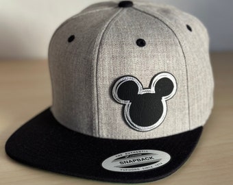 May include: A gray and black baseball cap with a black Mickey Mouse silhouette on the front. The cap has a black brim and a white label that reads "The Authentic Yupoong Snapback Since 1974".