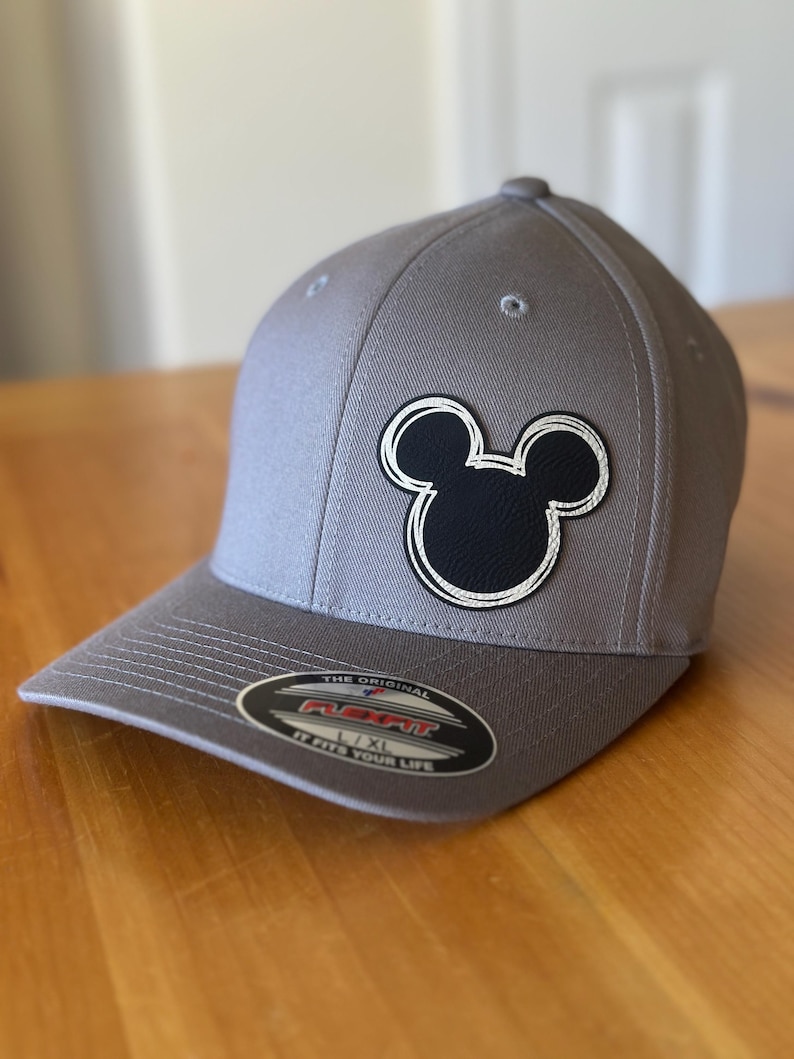 K&ouml;nnte beinhalten: Graue Baseballkappe mit einem schwarz-wei&szlig;en Mickey-Maus-Motiv. Die Kappe hat ein schwarzes und rotes "The Original Flexfit"-Etikett mit dem Text "L/XL It Fits Your Life".