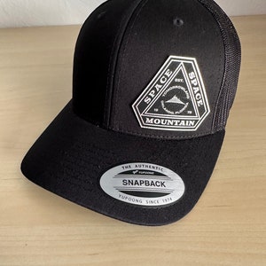 May include: Black snapback hat with a mesh back and a curved brim. The hat features a silver and black patch with the text "SPACE MOUNTAIN" and "TOMORROWLAND ORLANDO FLORIDA". The hat also has a "SNAPBACK" label.