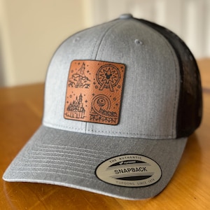 May include: A gray and black baseball cap with a brown leather patch featuring a black and white illustration of a Ferris wheel, castle, and other attractions. The cap has a black mesh back and a label that reads "The Authentic Yupoong Snapback Yupoong Since 1974."