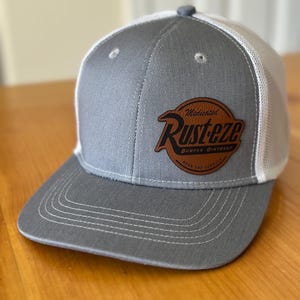 May include: Gray and white trucker hat with a brown leather patch that says "Medicated Rusteze Bumper Ointment.  Beach End Formula."