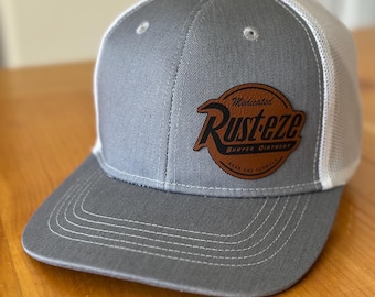May include: Gray and white trucker hat with a brown leather patch that says "Medicated Rusteze Bumper Ointment.  Beach End Formula."