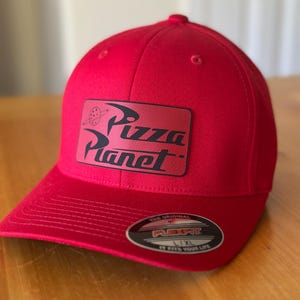 K&ouml;nnte beinhalten: Rote Baseballkappe mit einem schwarzen und wei&szlig;en "Pizza Planet" Logo-Patch auf der Vorderseite. Die Kappe hat ein Flexfit-Etikett auf der R&uuml;ckseite.