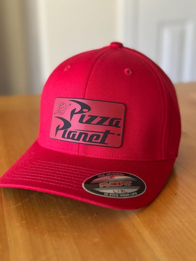 K&ouml;nnte beinhalten: Rote Baseballkappe mit einem schwarzen und wei&szlig;en "Pizza Planet" Logo-Patch auf der Vorderseite. Die Kappe hat ein Flexfit-Etikett auf der R&uuml;ckseite.