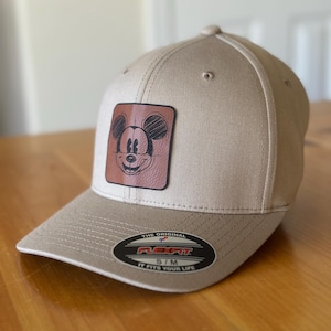 K&ouml;nnte beinhalten: Eine beige Baseballkappe mit einem braunen Lederpatch, auf dem eine Skizze von Mickey Mouse zu sehen ist. Der Patch befindet sich in der Mitte der Kappe. Die Kappe hat ein Flexfit-Etikett mit dem Text "The Original Flexfit S/M It Fits Your Life".