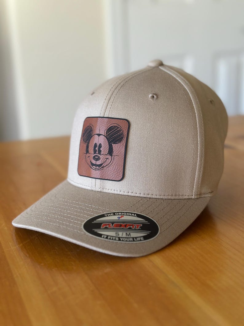 K&ouml;nnte beinhalten: Eine beige Baseballkappe mit einem braunen Lederpatch, auf dem eine Skizze von Mickey Mouse zu sehen ist. Der Patch befindet sich in der Mitte der Kappe. Die Kappe hat ein Flexfit-Etikett mit dem Text "The Original Flexfit S/M It Fits Your Life".