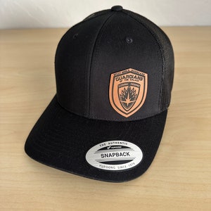 May include: Black and brown Guardians of the Galaxy themed baseball cap with a mesh back. The cap has a brown leather patch with the Guardians of the Galaxy logo and the text "EPCOT WORLD SHOWCASE" and "COSMIC REIGN." The cap also has a silver sticker with the text "THE AUTHENTIC YUPOONG SNAPBACK YUPOONG SINCE 1974."