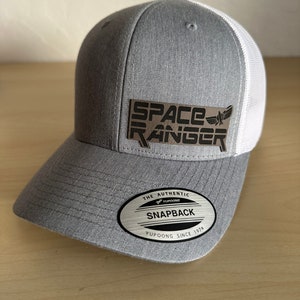 May include: A gray and white baseball cap with a silver patch that says "SPACE RANGER" in black letters. The cap has a white mesh back and a gray front. The cap has a black and silver sticker that says "THE AUTHENTIC SNAPBACK YUPOONG SINCE 1974".