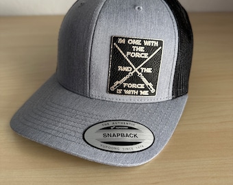 May include: A gray and black trucker hat with a black leather patch that says "I'm one with the force and the force is with me". The patch has two crossed lightsabers. The hat has a black mesh back and a gray front with a silver YUPOONG logo and the text "The Authentic Snapback YUPOONG Since 1974".