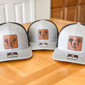 May include: Three gray and black trucker hats with a brown leather patch featuring a cartoon image of Mickey Mouse and Minnie Mouse. The hats are on a wooden surface.