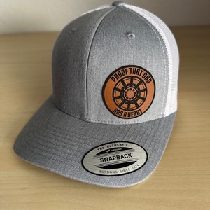 May include: Gray and white trucker hat with a brown leather patch that says "Proof That Dad Has A Heart". The patch features a stylized gear design. The hat has a silver oval label that says "The Authentic Yupoong Snapback Yupoong Since 1974".