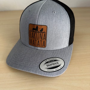 May include: A gray and black snapback hat with a leather patch featuring the words "Hakuna Matata" and silhouettes of a lion and a meerkat. The hat has a curved brim and a mesh back.