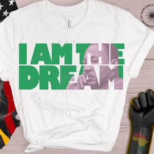 May include: White t-shirt with a green and pink graphic design that says "I AM THE DREAM" with a picture of Martin Luther King Jr. in the center.